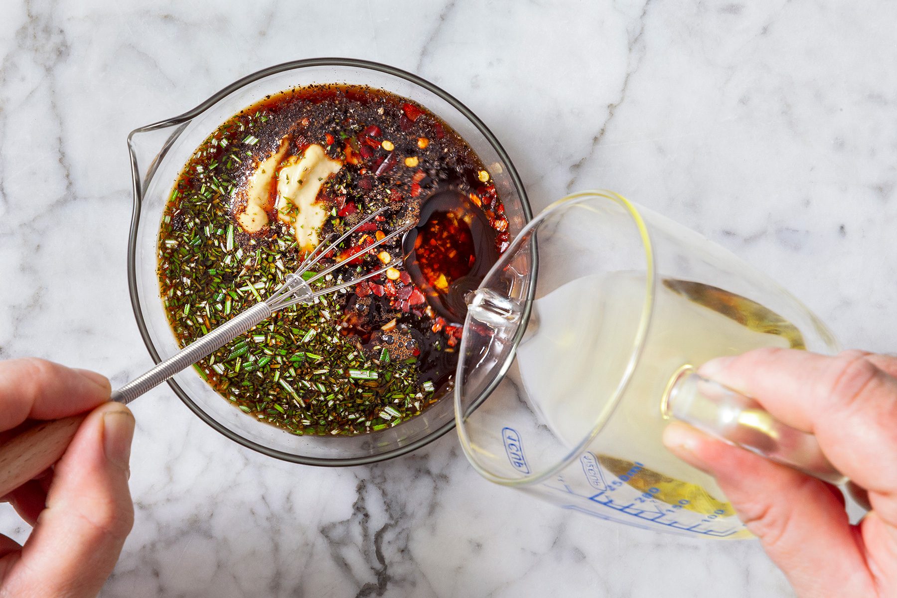 overhead shot of a vibrant marinade or dressing in a glass bowl atop a marble surface, ingredients including finely chopped herbs, red pepper flakes, and what appears to be melted butter or minced garlic, The mixture displays layers of golden oil, dark liquids resembling soy sauce or vinegar, and specks of various spices, A hand is shown whisking the concoction with a metal whisk, while another hand pours a clear liquid, likely oil or vinegar, from a measuring cup into the bowl,