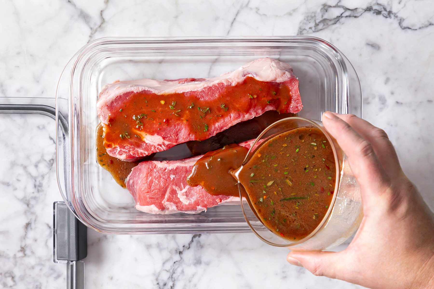 overhead shot of raw steaks in a clear glass container on a marble surface, A hand is pouring a rich, herb-flecked marinade over the meat from a glass measuring cup, The raw steaks are a vibrant pink color;