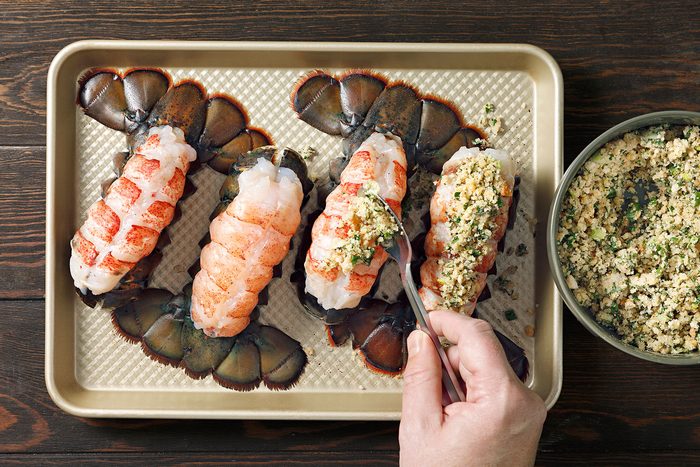 Hand preparing four lobster tails on a baking tray, applying a breadcrumb and herb mixture.
