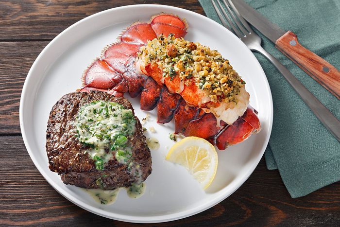 A white plate with a seasoned steak topped with herb butter and a baked lobster tail garnished with breadcrumbs. A lemon wedge is on the side, and silverware rests on a green napkin.