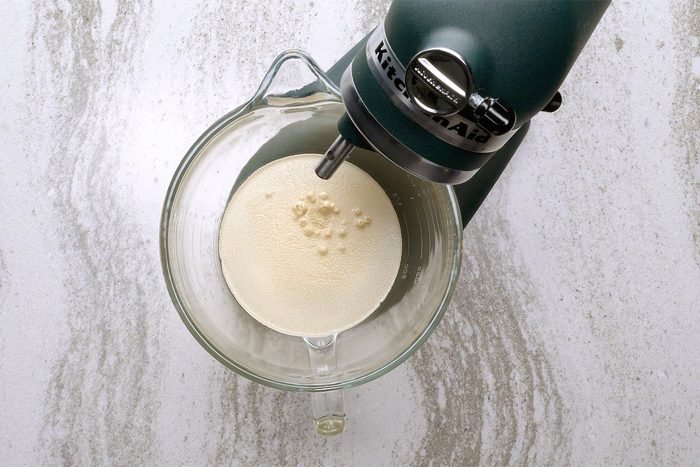 A top-down view of a stand mixer with a glass bowl attached. The bowl contains a light-colored creamy mixture, and the mixer is set on a marble countertop.