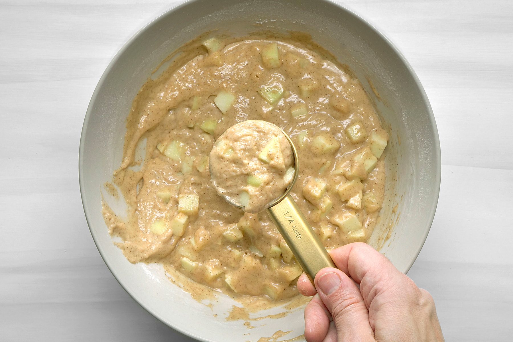 overhead shot of a bowl of batter with diced apples in it; a hand is holding a measuring cup that has some batter on it; the bowl and measuring cup are on a white surface