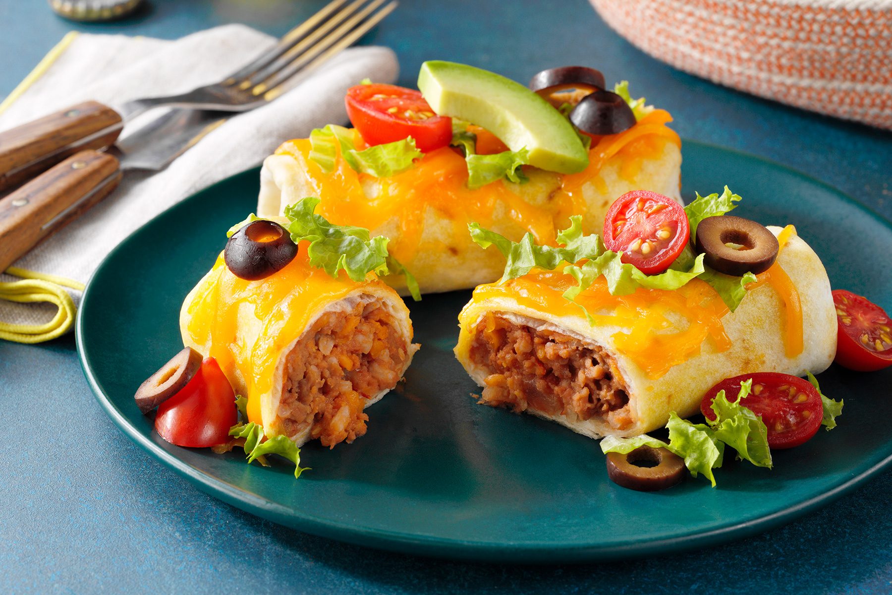 Two burritos topped with melted cheese, sliced black olives, and cherry tomatoes on a green plate. The burritos are garnished with avocado slices and lettuce, and one is cut open to reveal a filling of beans and rice. A fork and knife rest nearby.