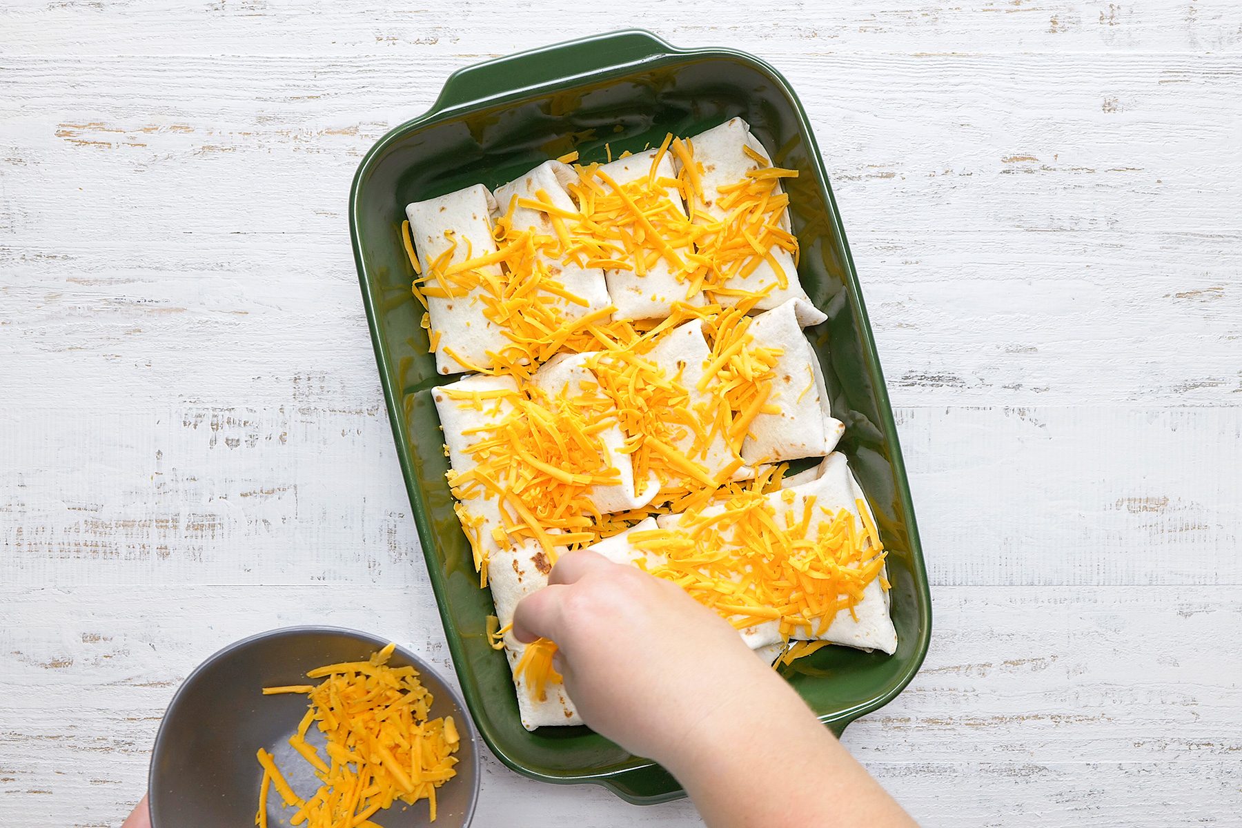A hand is sprinkling shredded cheddar cheese over six burritos in a green baking dish. The background is a white wooden surface, and there is a small bowl with more cheese in the lower corner.