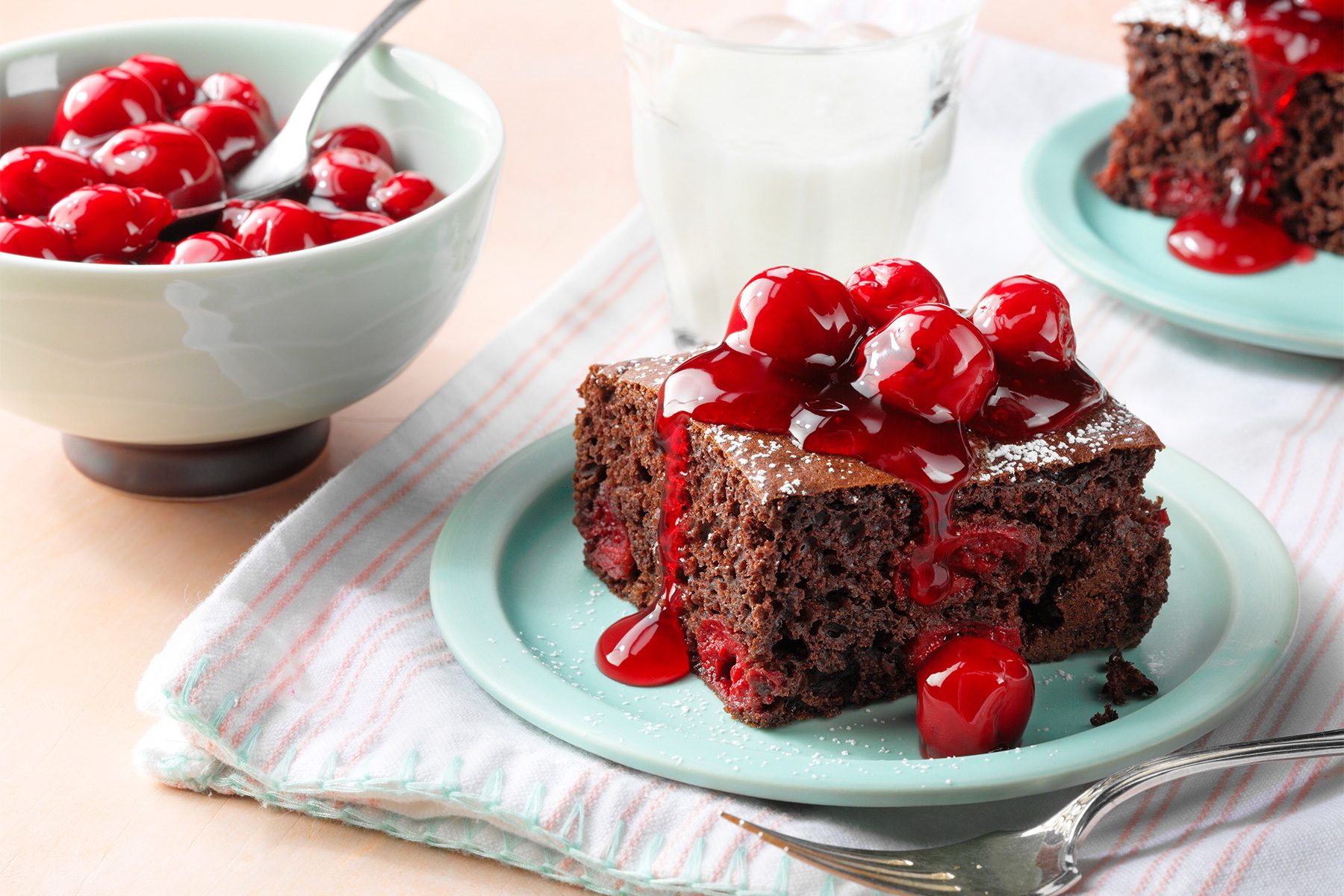 A slice of chocolate cake topped with glazed cherries on a light blue plate. Next to it is a bowl of cherries and a glass of milk. A fork rests on the striped napkin under the plate.