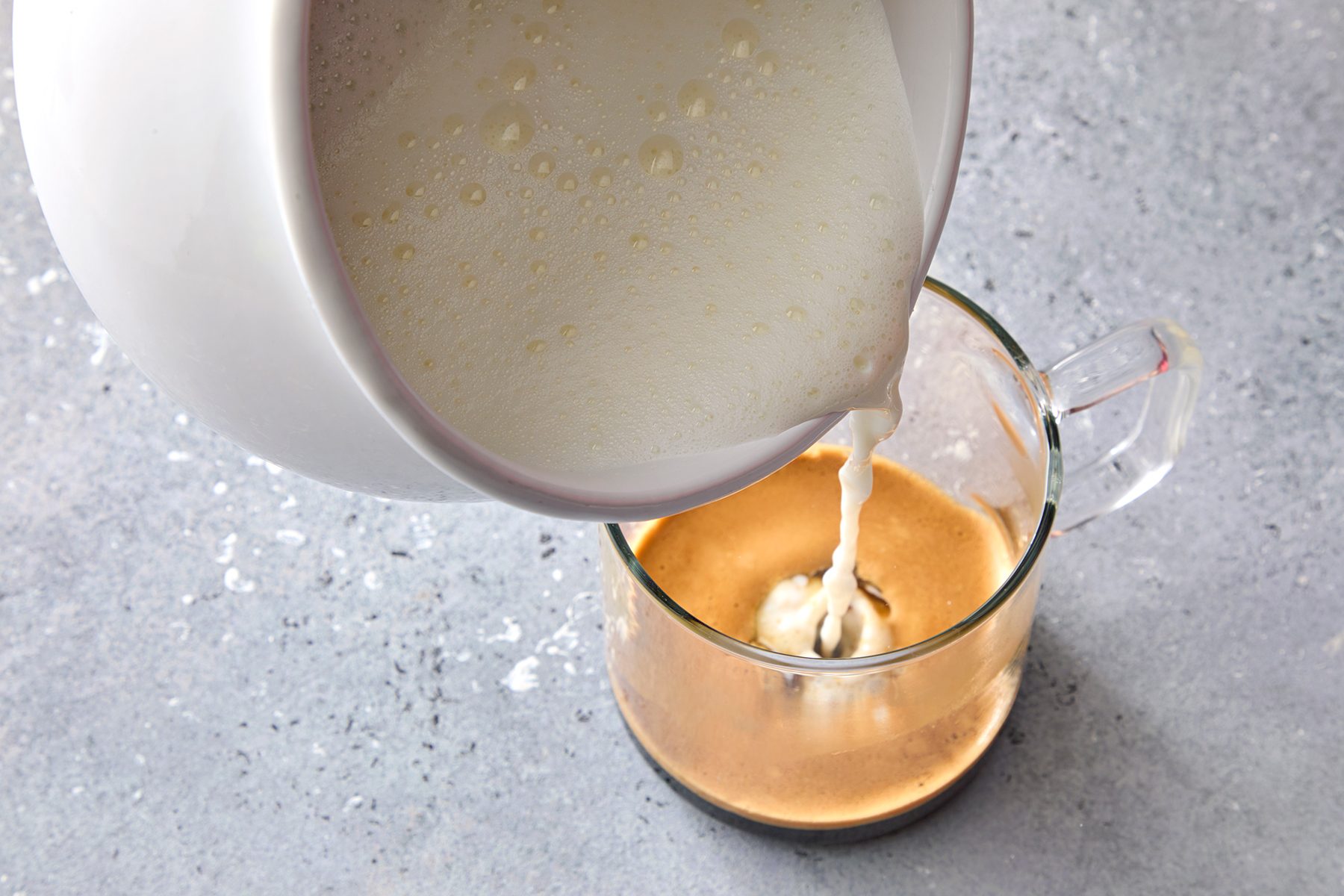 A white pitcher pours frothy milk into a glass mug containing espresso on a textured gray surface, creating a creamy latte.