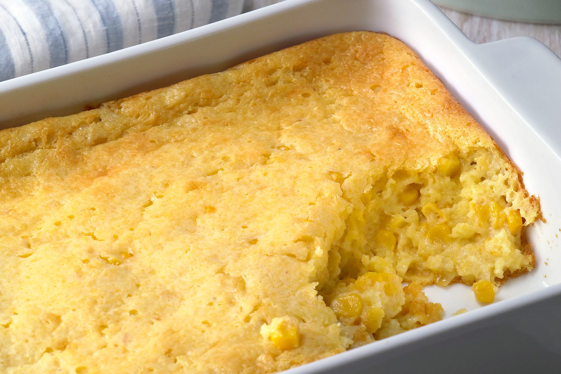 3/4th shot of a casserole dish filled with cornbread pudding, a blue and white striped kitchen towel beside it