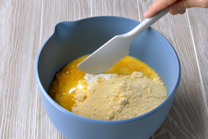 3/4th shot of a blue mixing bowl on a wooden surface, containing a mixture of ingredients for a recipe; the bowl is filled with yellow beaten eggs, a white spatula, held by a hand, is used to combine the ingredients