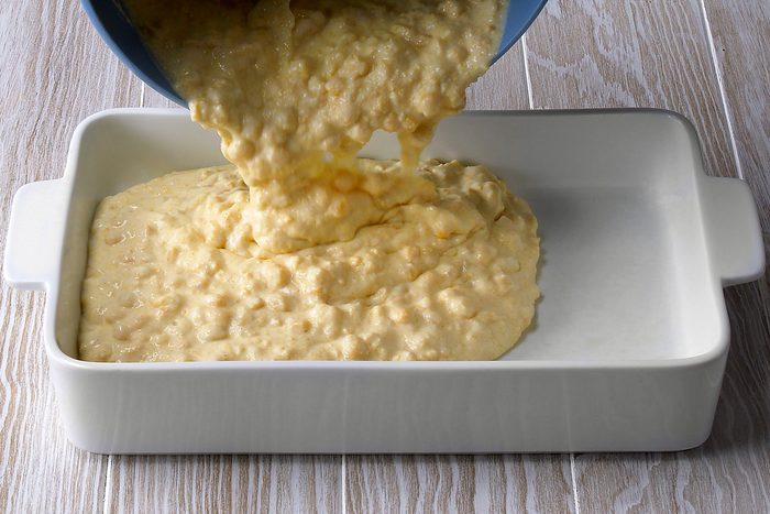 3/4th shot of a white ceramic baking dish sitting on a light wooden surface; a blue bowl is being tilted over the baking dish, pouring a thick, yellow, creamy substance into it;