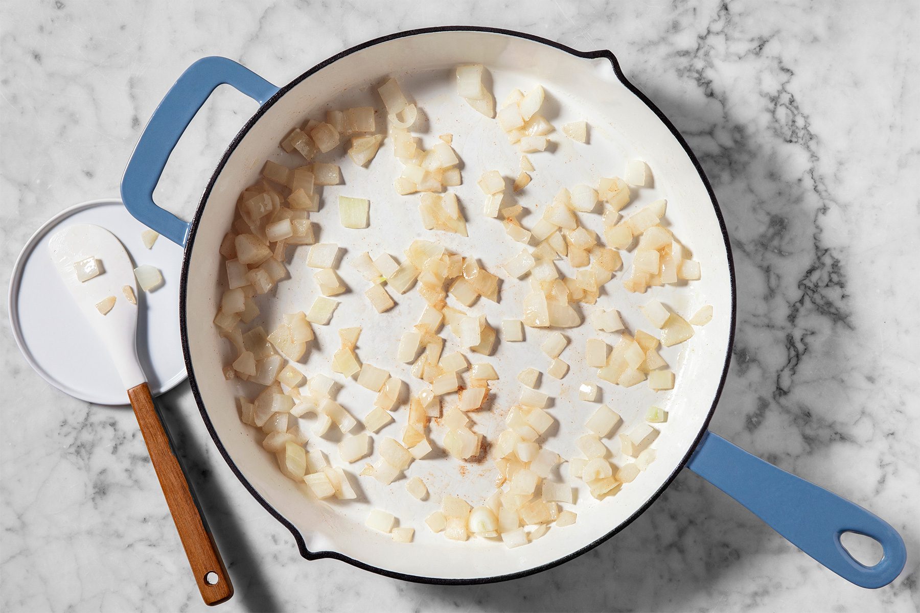 A white frying pan with a blue handle sits on a marble countertop. Inside the pan are evenly spread, small diced pieces of onions. A wooden-handled spatula with some onion pieces is next to the pan.