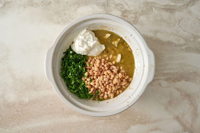 Overhead view of navy beans, sour cream, and fresh cilantro in the slow cooker with shredded chicken for the Taste of Home Green Chicken Chili recipe.
