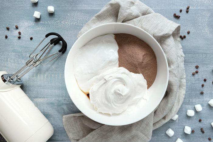 A bowl with whipped cream and chocolate mousse sits on a textured blue surface, surrounded by scattered mini marshmallows and chocolate chips. An electric hand mixer with beaters is nearby, resting on a light gray cloth.