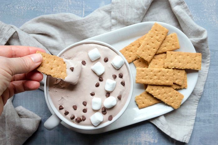 A person dips a graham cracker into a bowl of chocolate dip, topped with marshmallows and chocolate chips. The bowl is on a white platter with more graham crackers, set on a textured gray surface with a beige cloth.