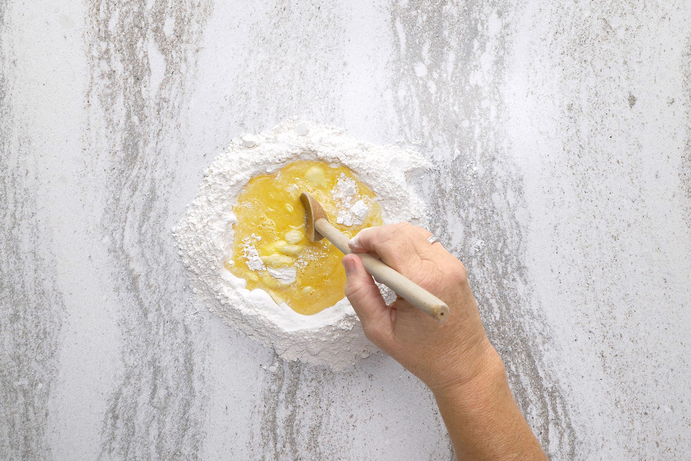 Eggs and water in the centre of flour mix with a hand holding a wooden ladle