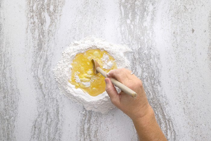 Eggs and water in the centre of flour mix with a hand holding a wooden ladle