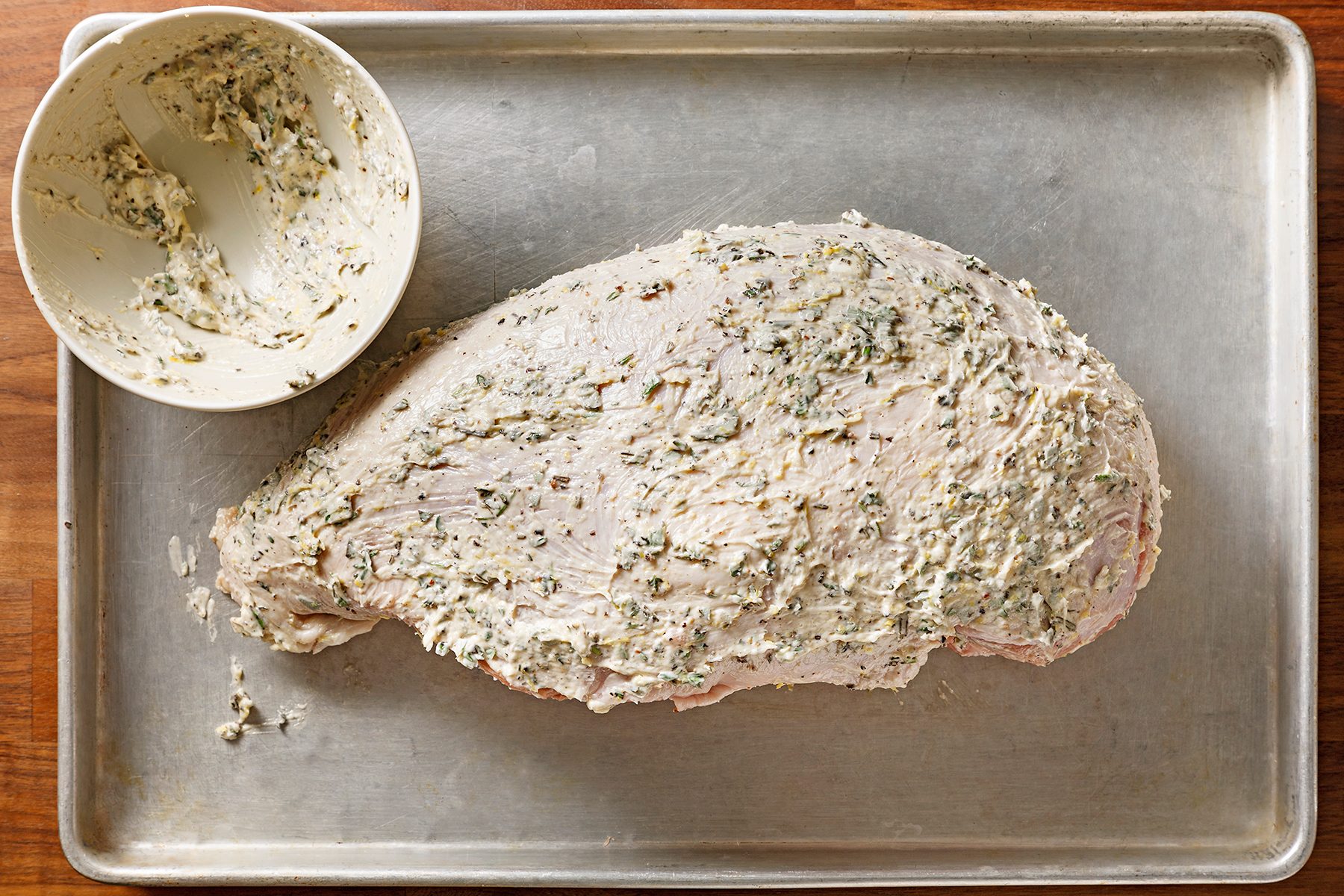 A large piece of meat coated with a herb-infused cream mixture is on a metal baking tray. A bowl containing remnants of the coating mixture is placed on the tray's corner. The tray is on a wooden surface.