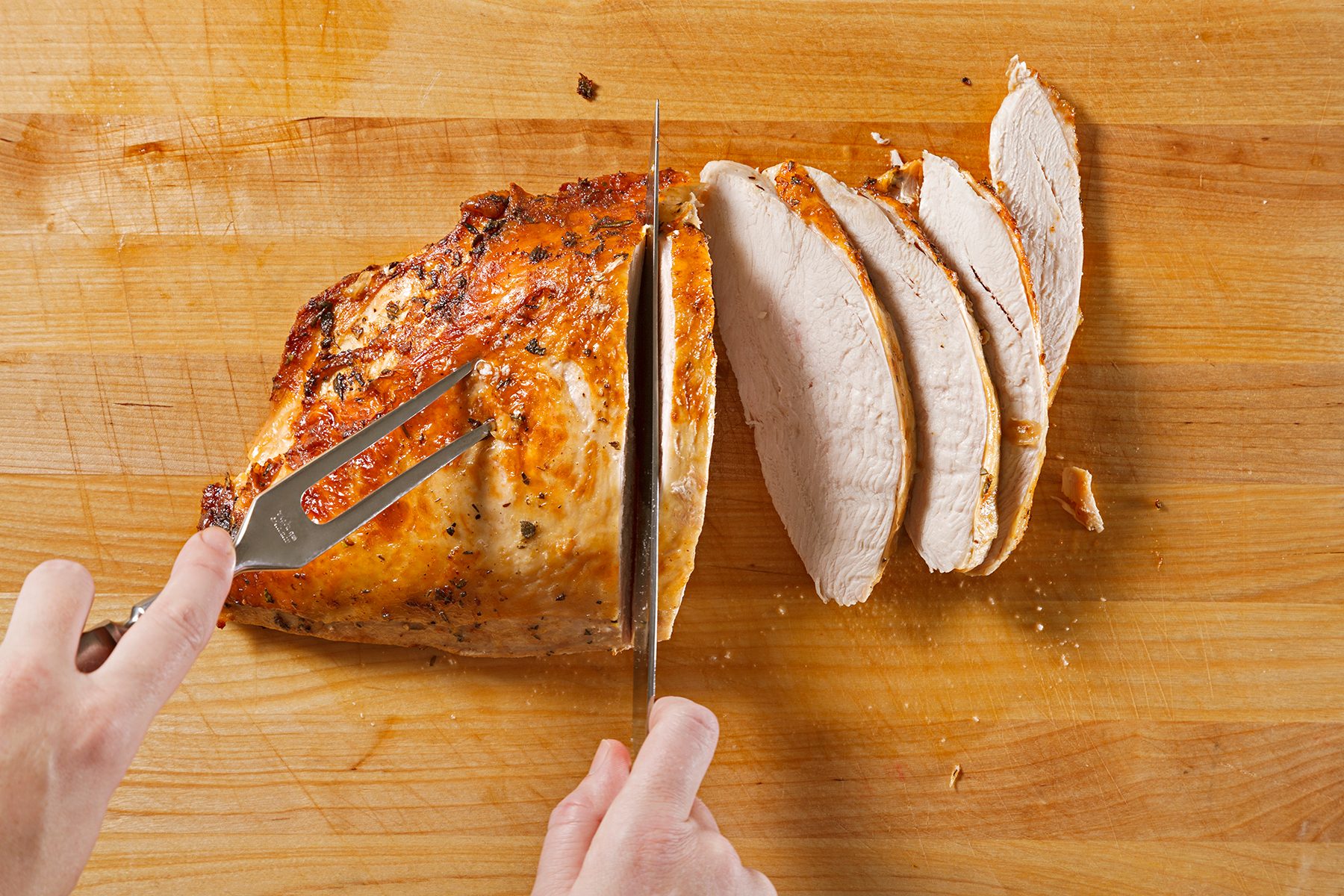 A person slices a cooked, golden-brown piece of turkey breast on a wooden cutting board, revealing several juicy, evenly cut slices. A fork secures the meat while a knife carves through it.