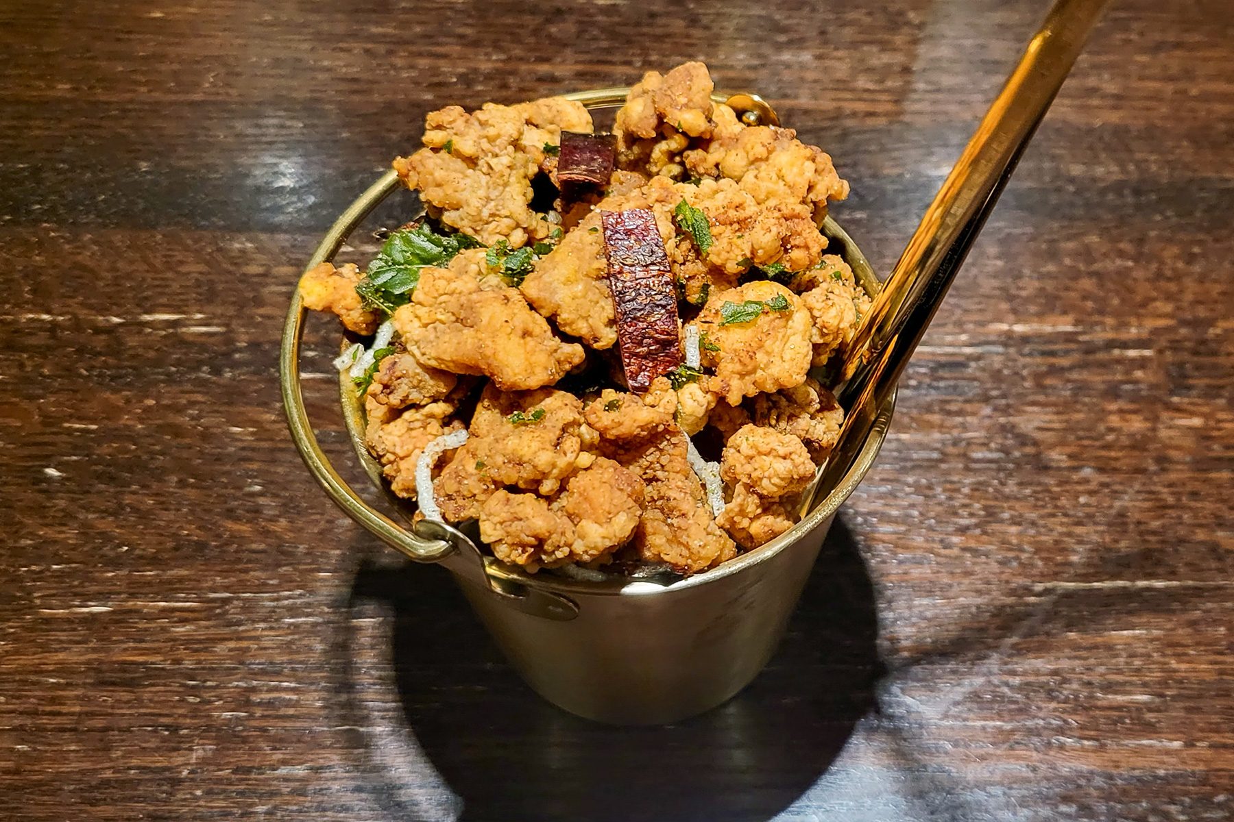 A metal cup filled with crispy fried chicken pieces garnished with herbs, placed on a dark wooden table. A shiny golden spoon is inserted into the cup.