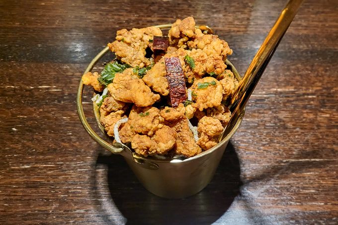 A metal cup filled with crispy fried chicken pieces garnished with herbs, placed on a dark wooden table. A shiny golden spoon is inserted into the cup.