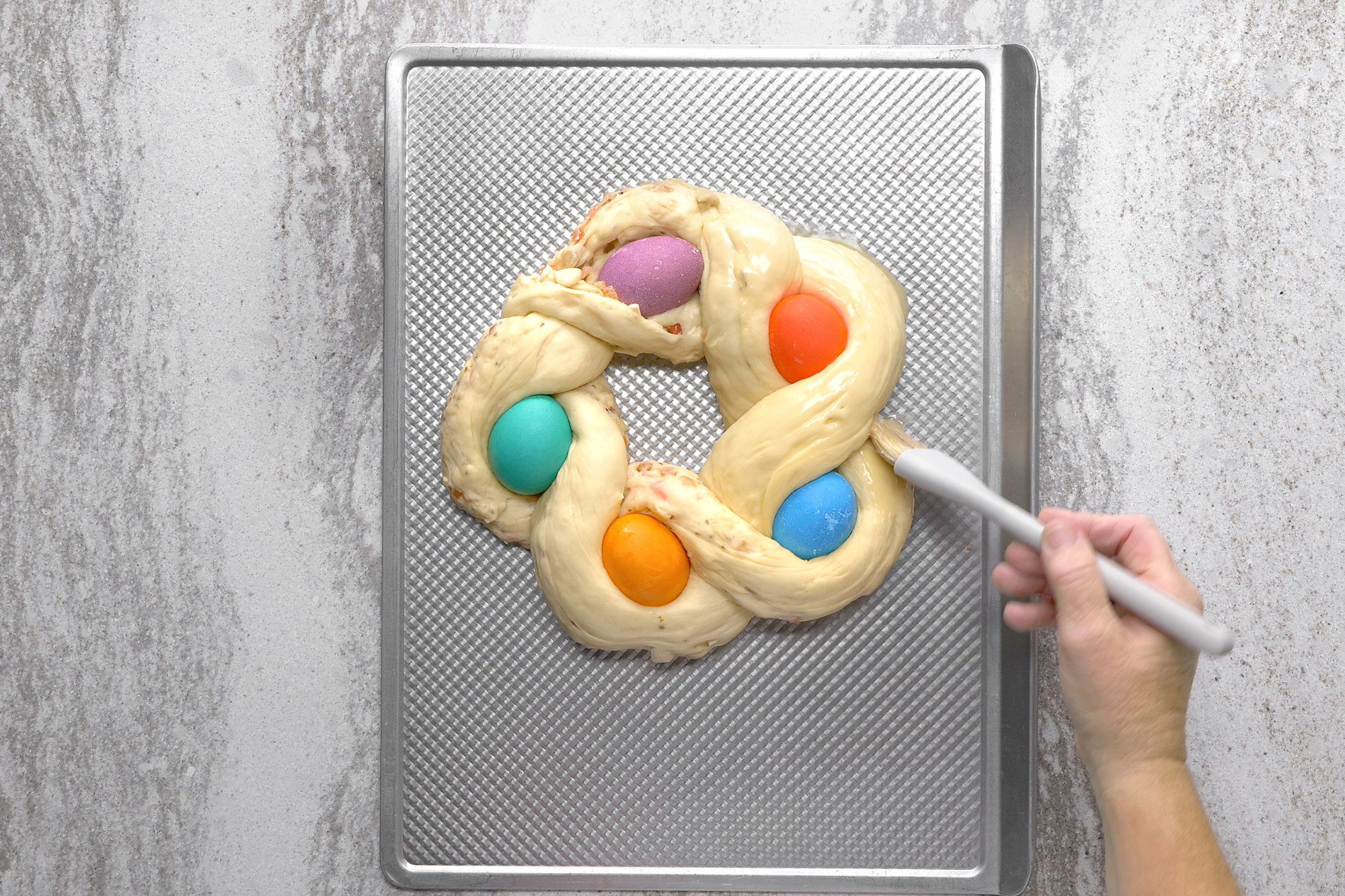 overhead shot of A hand with a pastry brush is brushing the top of a braided Easter bread on a baking sheet; the bread is shaped like a ring and has five colorful Easter eggs embedded in it; the baking sheet is on a white counter top; the bread is golden brown and shiny