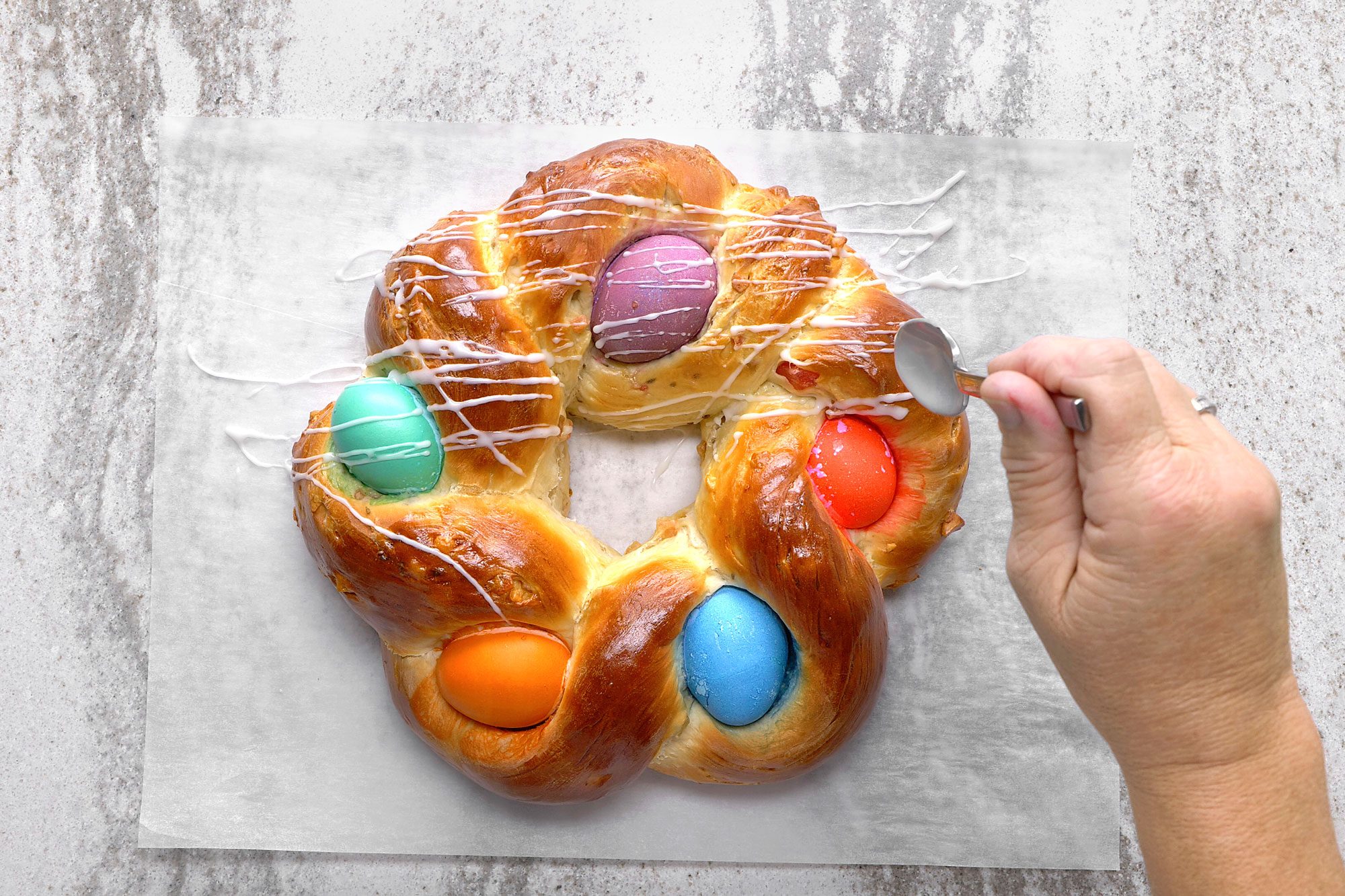 overhead shot of a hand is drizzling a white glaze over a homemade easter bread ring; the bread is decorated with colorful eggs, and the glaze is being drizzled over the top, the bread is sitting on a piece of parchment paper, and the background is a light gray countertop