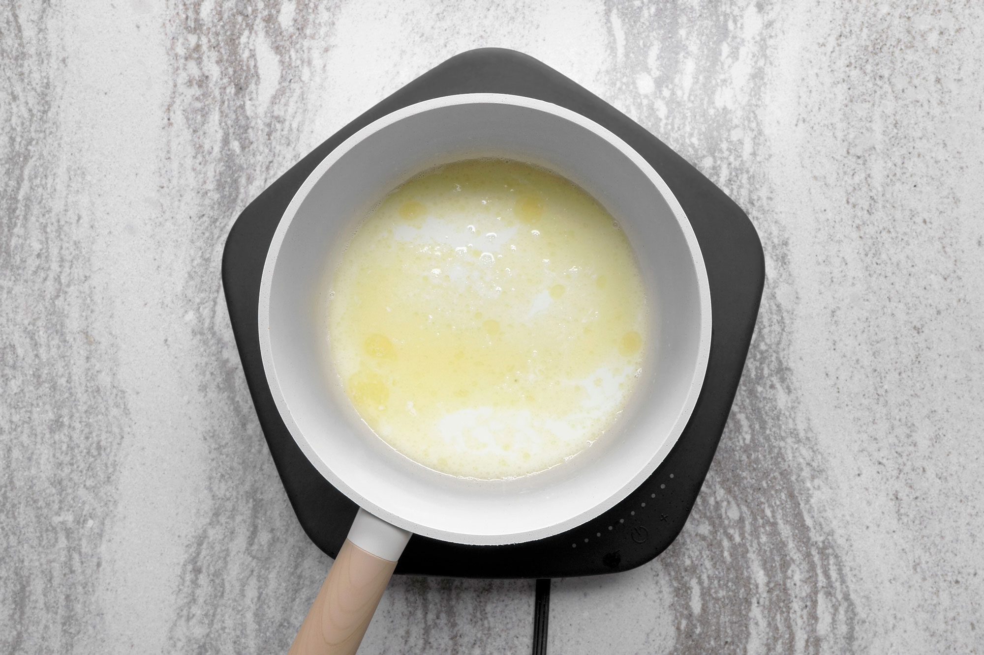 overhead shot of a white pot with melted butter sits on a black electric stovetop that is turned on, the stovetop is on a gray countertop; the pot has a wooden handle and the stovetop has a cord hanging down