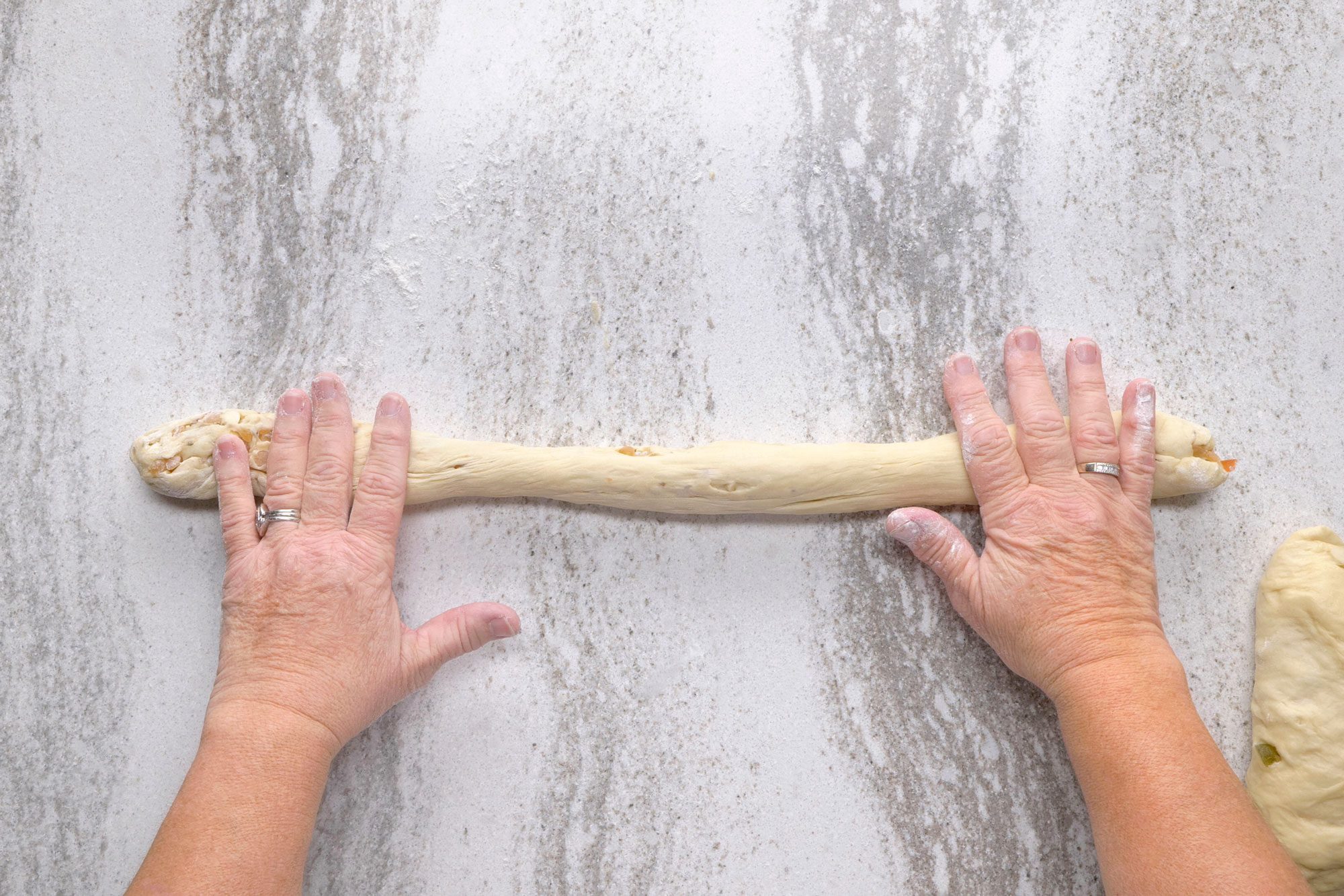 overhead shot of a person's hands are shown rolling a long piece of dough on a counter; they are pressing the dough down with their fingers to create a long, even shape; a ring is visible on their left ring finger; to the right of the dough there is another piece of dough which appears to be a single, round roll; the countertop is a light gray color
