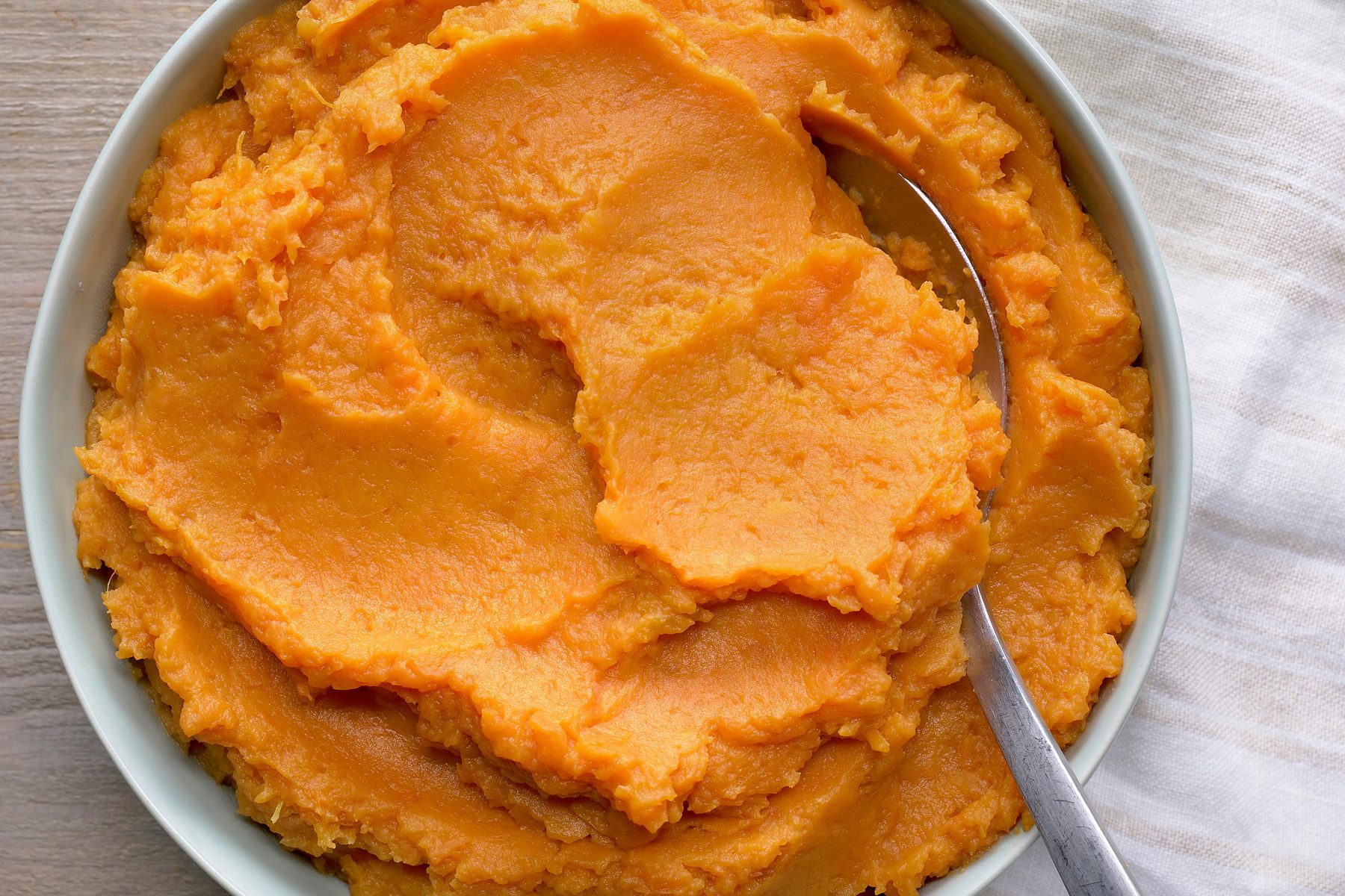overhead shot of a bowl of mashed sweet potatoes, with a spoon resting on top; the sweet potatoes are a vibrant orange color, and the bowl is a pale blue-green;