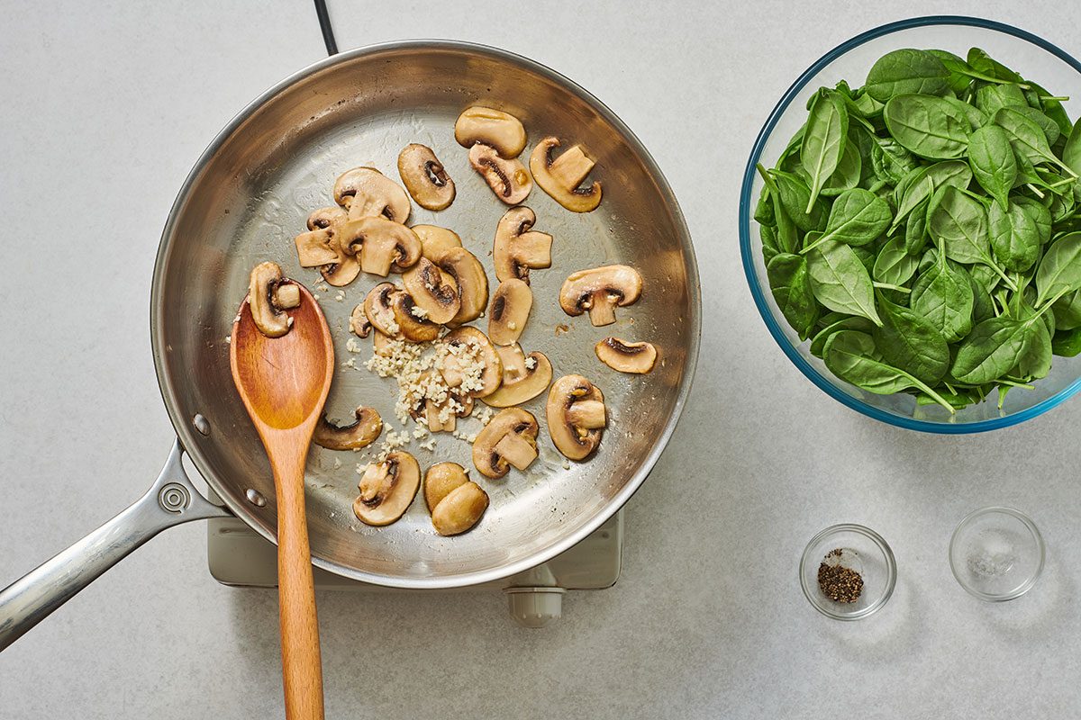 Sautéed mushrooms in a large skillet with a bowl of spinach nearby