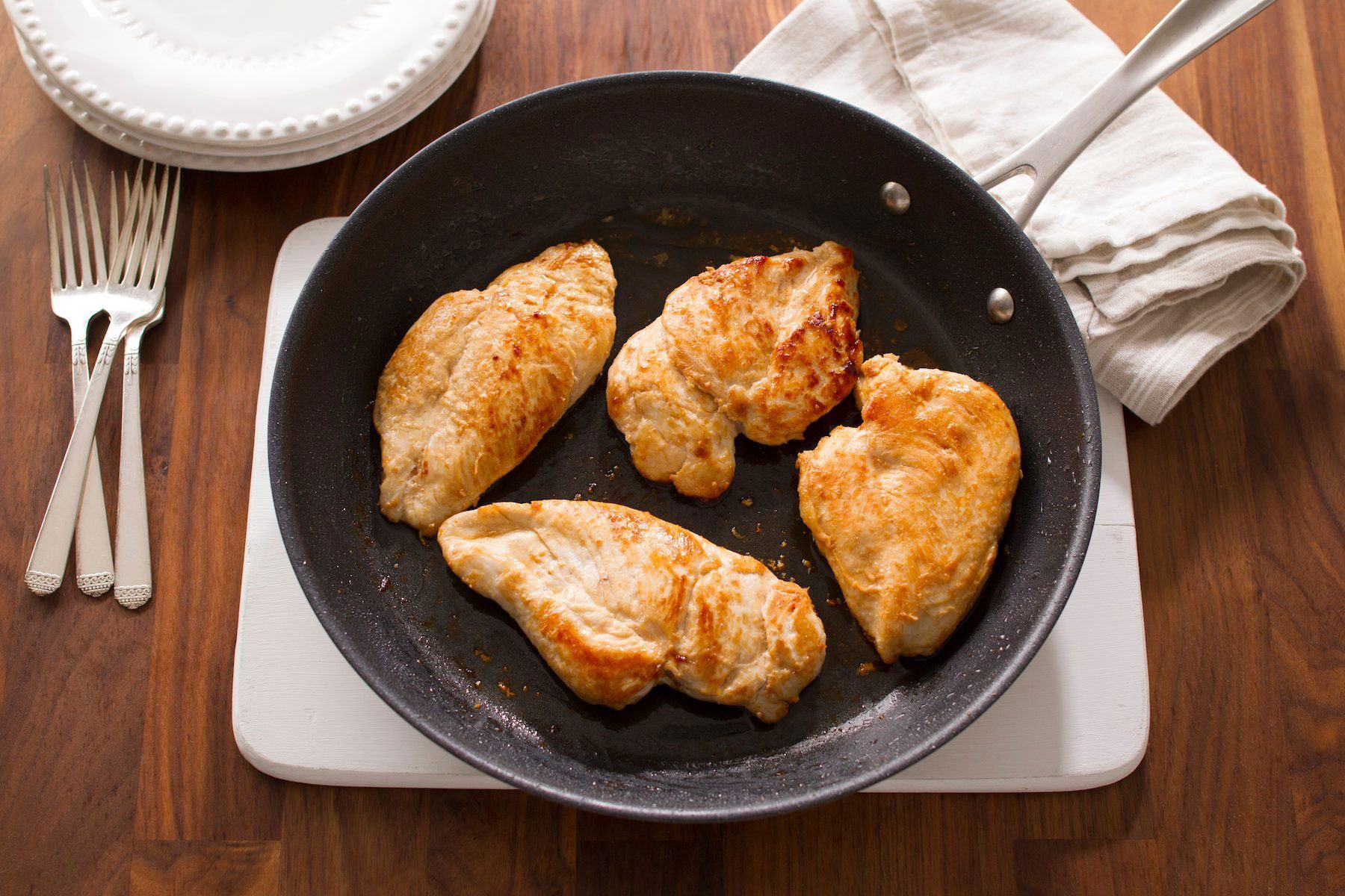 A pan with four cooked chicken breasts on a wooden surface. Nearby are a stack of white plates, a white cloth, and three forks.