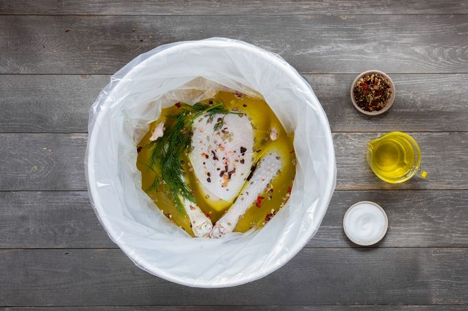 Raw turkey breast and drumsticks marinating in a container with oil, herbs, and spices. A small bowl of mixed spices, a bowl of olive oil, and a bowl of salt are placed beside the container on a wooden surface.