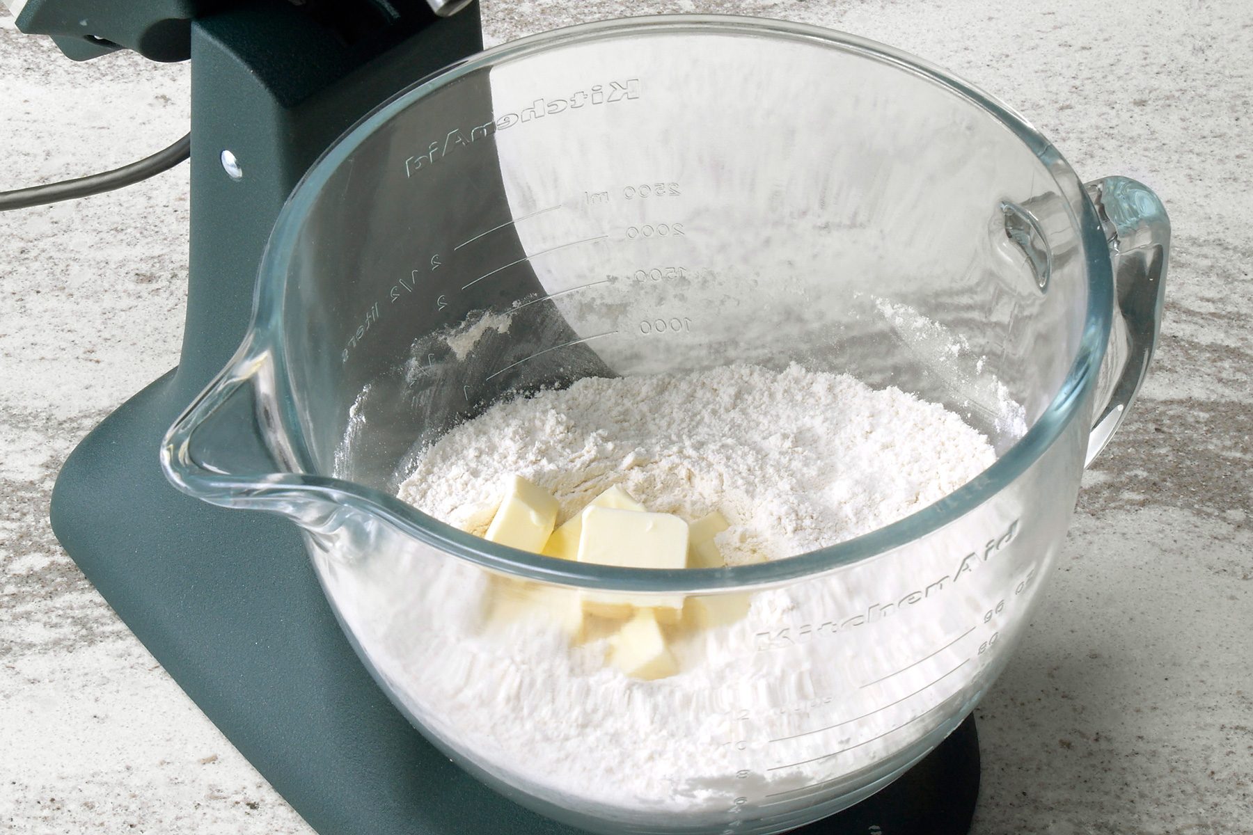 a close-up view of a kitchenaid stand mixer bowl; the bowl is made of glass and has a handle on the right side; the bowl is filled with white flour and a few chunks of butter; the bowl has markings on the side showing measurements in cups and milliliters; the bowl sits on a counter top that is speckled white and gray