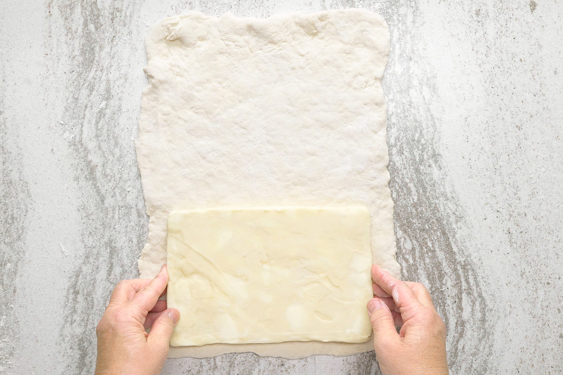 overheda shot of a piece of dough on a floured surface, a hand is placing a square of butter onto the dough