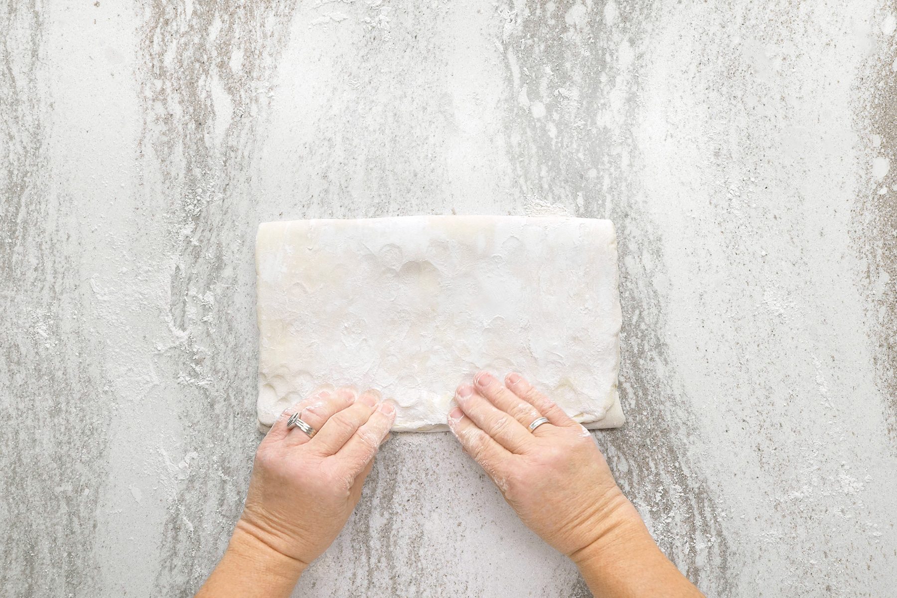 overhead shot of a person's hands holding a sheet of dough, the dough is lightly dusted with flour and the person is in the process of folding the dough in half, the background is a light grey surface with a white speckles