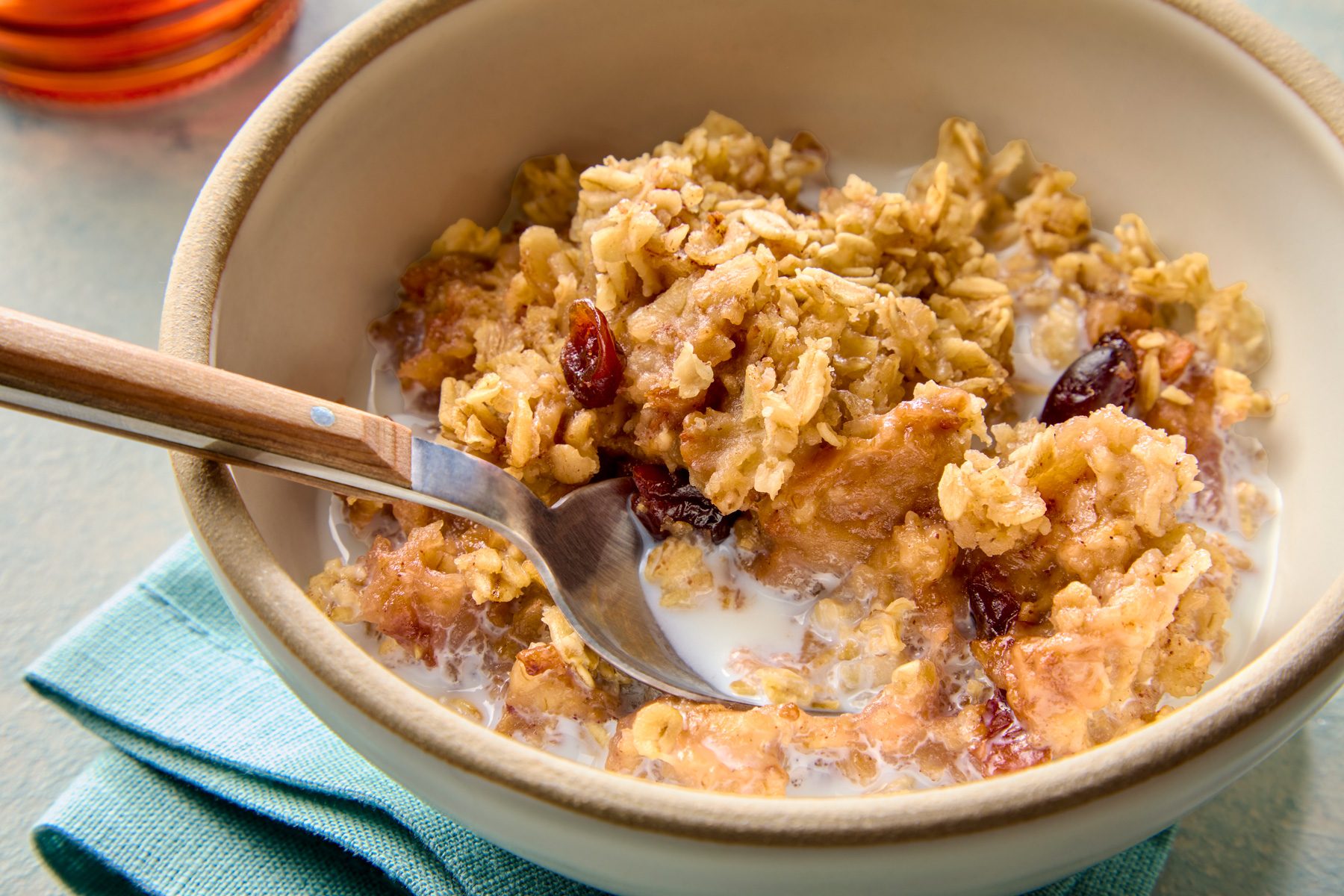 3/4th shot of a bowl of oatmeal with milk, topped with a layer of baked apples and cranberries; the bowl is sitting on top of a blue napkin, and there is a wooden spoon next to the bowl; and it is topped with a thick layer of milk