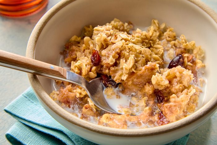 3/4th shot of a bowl of oatmeal with milk, topped with a layer of baked apples and cranberries; the bowl is sitting on top of a blue napkin, and there is a wooden spoon next to the bowl; and it is topped with a thick layer of milk