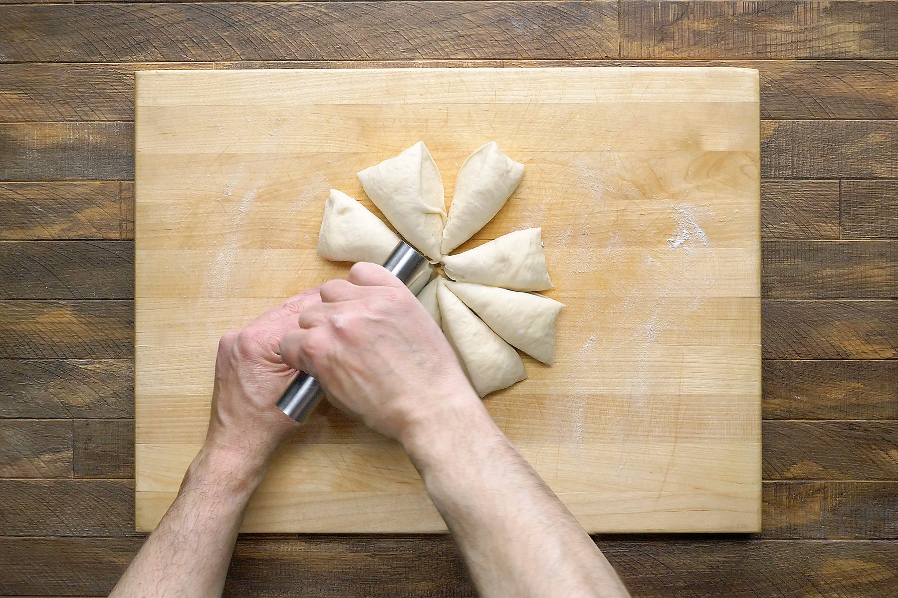 Hands holding a tool, cutting a round piece of dough into equal triangular sections on a wooden cutting board.