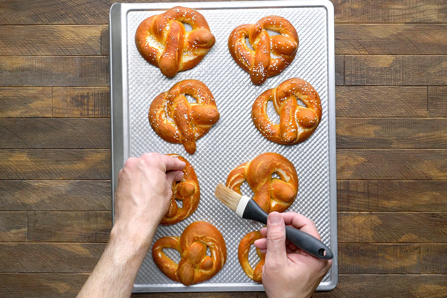 A person brushes melted butter on freshly baked pretzels on a silver baking tray. The tray rests on a wooden surface, and there are six pretzels visible.