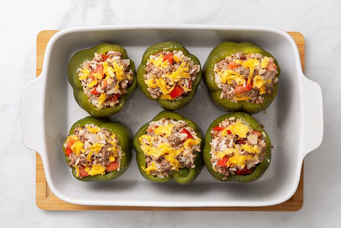 Green peppers stuffed with ground beef, tomatoes, rice, onion, and shredded cheddar cheese in a baking dish for step three of Stuffed Peppers with Cheese recipe for Taste of Home