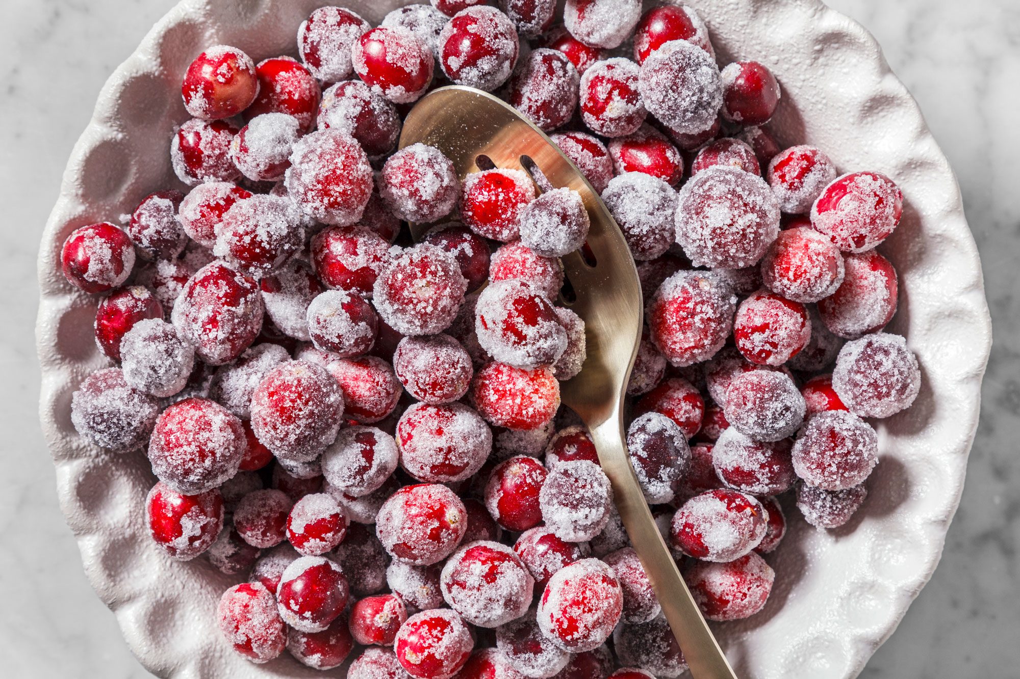 A white plate filled with sugared cranberries, covered in a dusting of sugar. A silver spoon rests among the berries.