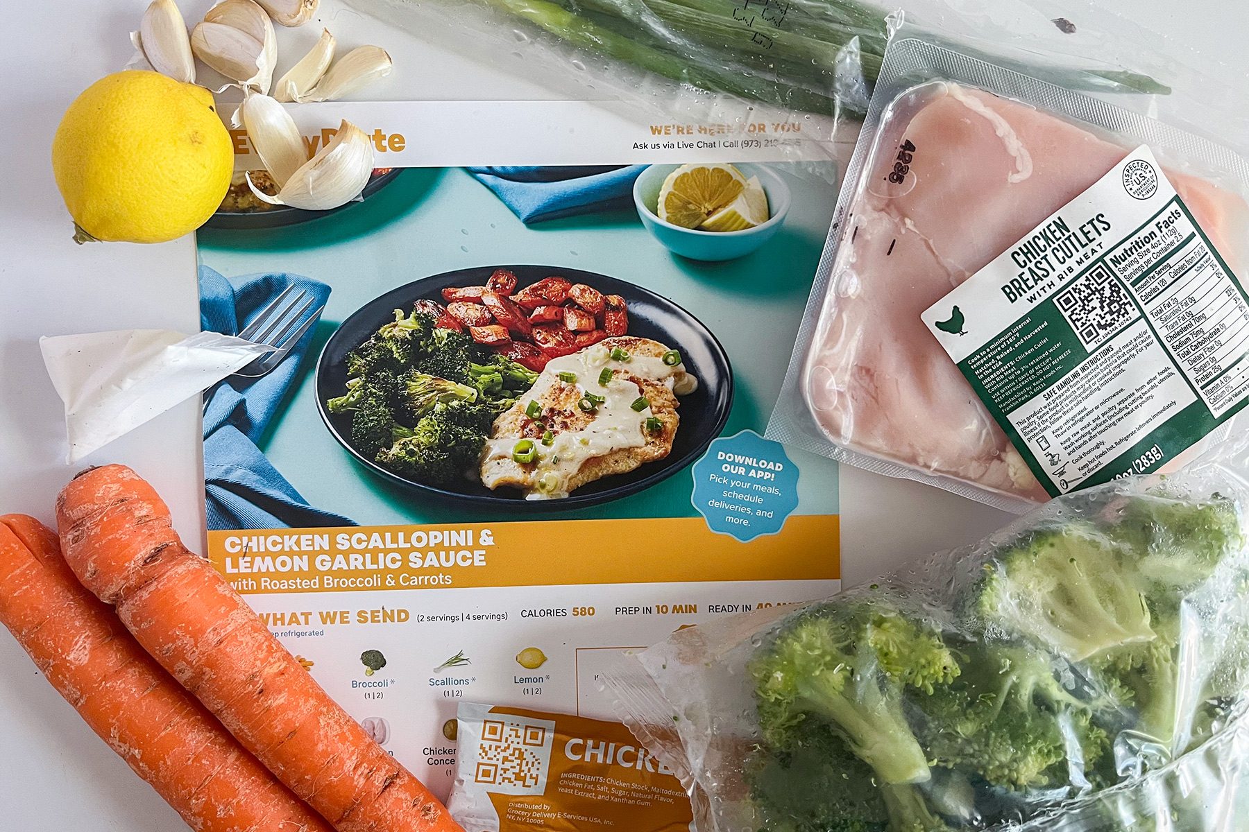 A meal kit featuring ingredients for chicken with scallion and lemon garlic sauce. Visible items include broccoli, carrots, chicken breast, scallions, garlic, a lemon, and a recipe card with a photo of the finished dish.