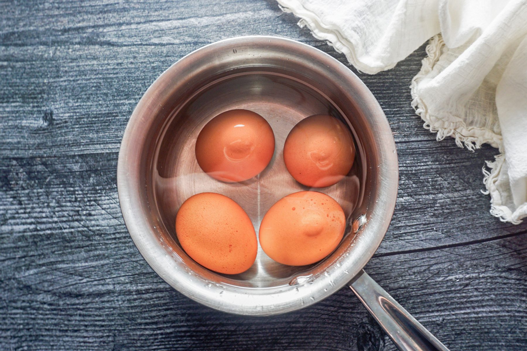 A top-down view of four brown eggs submerged in water inside a stainless steel pot placed on a dark wooden surface. A white cloth napkin is partially visible in the top right corner.