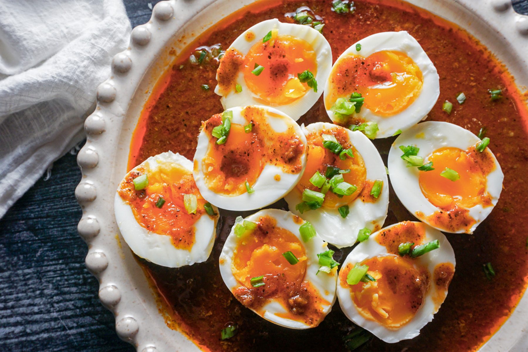 A plate of halved boiled eggs with runny yolks, topped with chopped green onions and drizzled with a spicy red sauce, set on a textured ceramic dish. A white napkin is partially visible on the side.