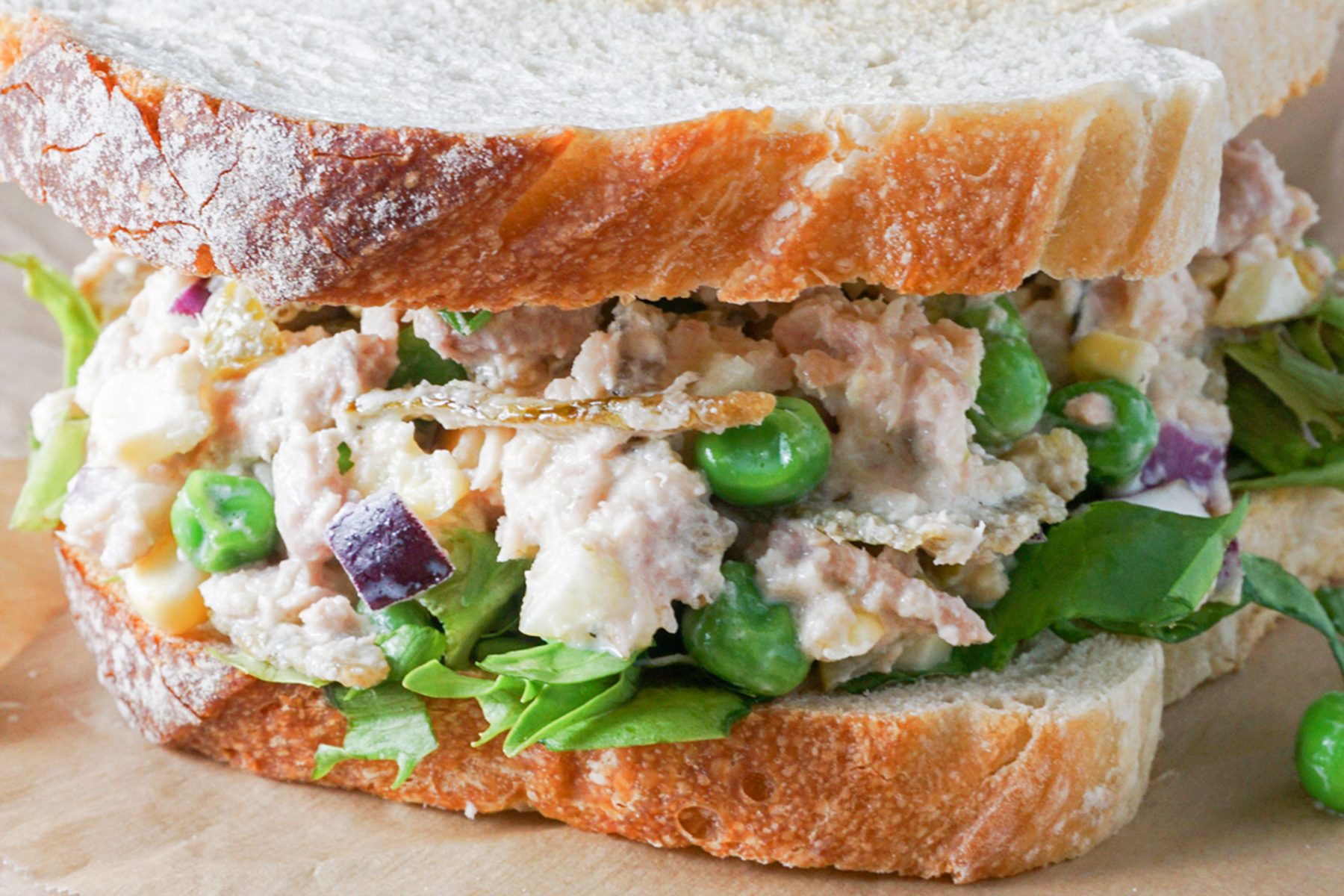 A close-up of a sandwich filled with a creamy tuna salad mixture, including tuna chunks, green peas, red onions, and leafy greens, between two slices of crusty bread.