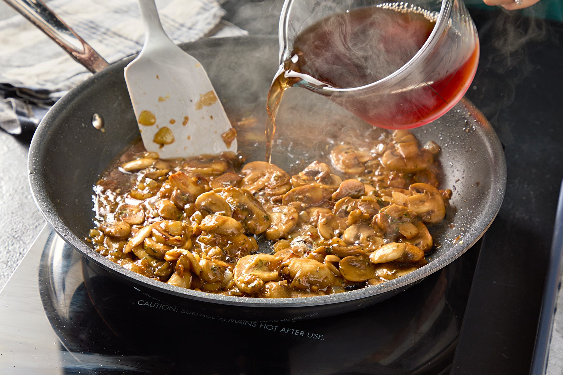 A person is pouring a brown liquid from a glass measuring cup into a skillet filled with cooked mushrooms and herbs on a stovetop. A white spatula is resting in the skillet.