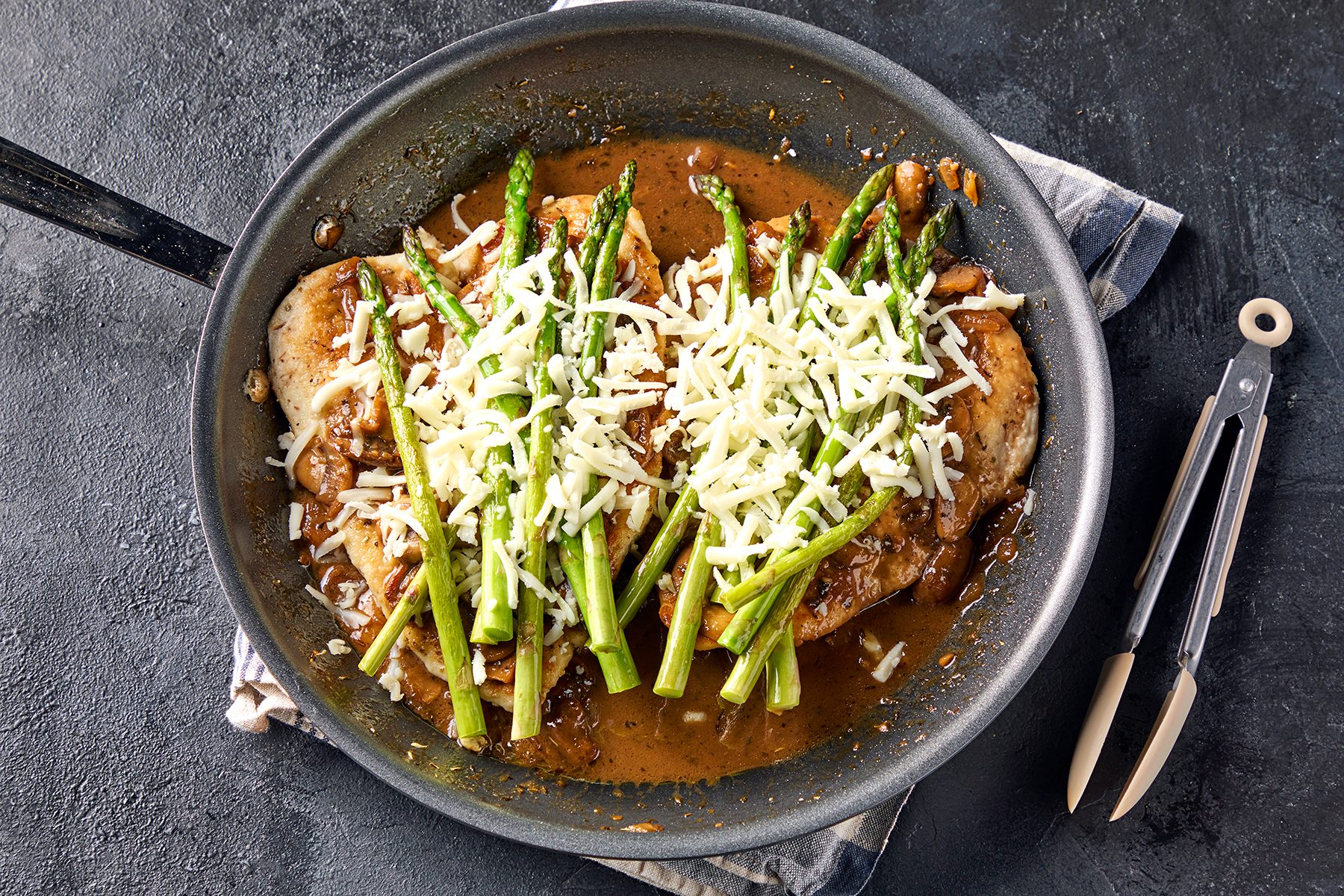 A skillet containing chicken breasts topped with shredded cheese and asparagus, set on a dark surface with a pair of serving tongs beside it. The dish is cooked and covered in a savory sauce.
