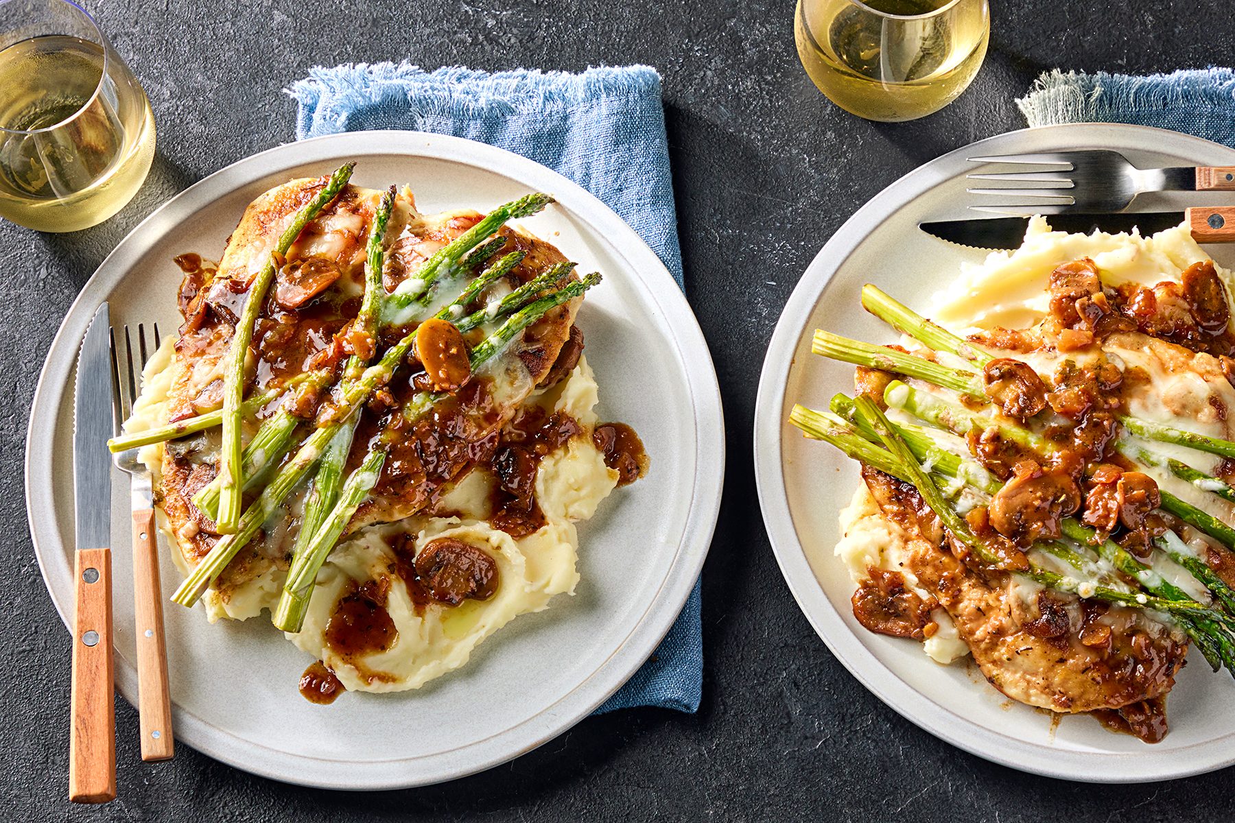 Two plates of mashed potatoes topped with chicken breast, asparagus, and a mushroom sauce. Wooden-handled cutlery lies beside each plate. Two glasses of white wine are in the background, with a blue cloth under one plate.