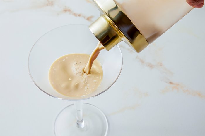 A person pours a beige creamy liquid from a shaker into a martini glass on a marble surface.