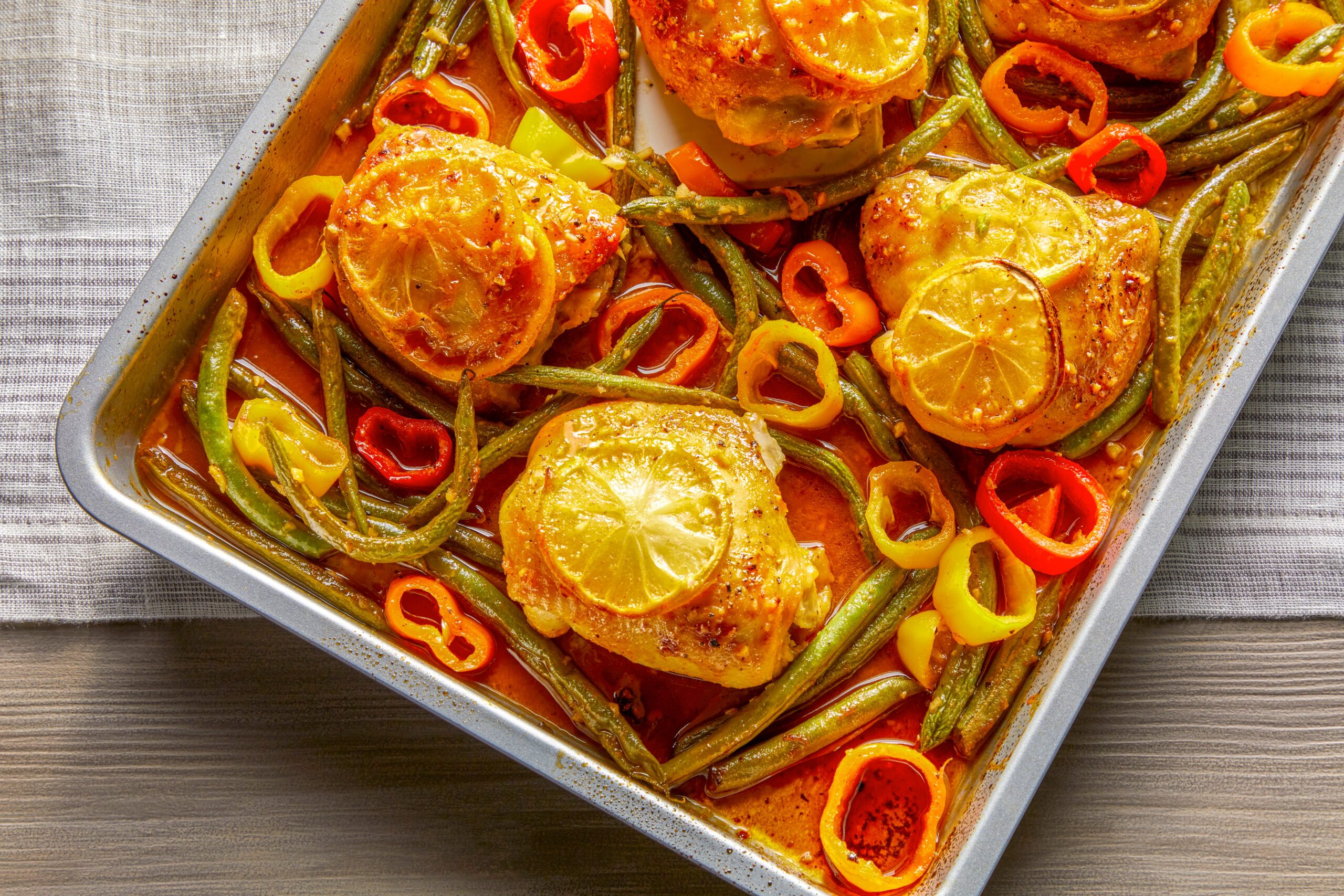 A sheet pan with baked chicken thighs topped with lemon slices, surrounded by green beans and sliced colorful bell peppers, sitting on a gray striped cloth. The dish appears flavorful and vibrant.