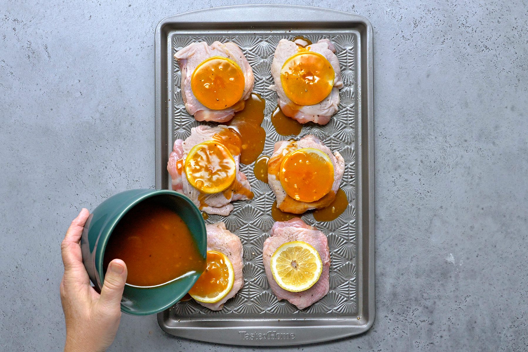 A hand pouring orange sauce from a green bowl onto slices of cooked meat arranged on a baking tray. Each meat slice is topped with an orange slice and a lemon slice. The tray rests on a gray countertop.